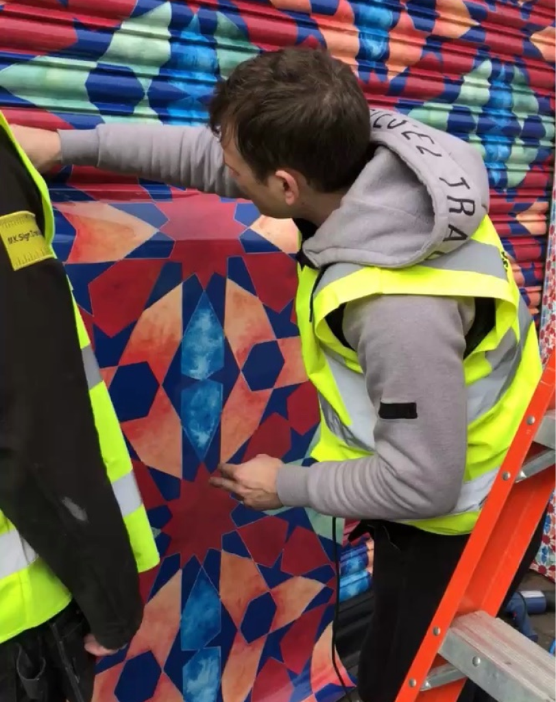 Close-up of a worker in a yellow safety vest carefully trimming or adjusting a vinyl graphic on a metal surface.