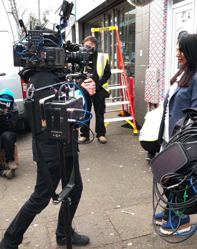 A film crew with a large professional camera filming the installation process of a storefront vinyl wrap on a city street.