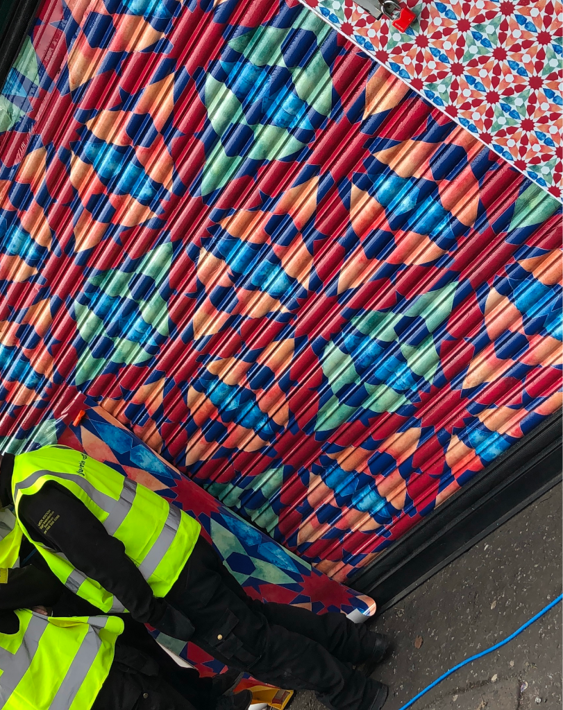 Close-up of a vibrant, geometric vinyl wrap featuring blue, red, and yellow patterns being applied to a corrugated metal storefront shutter.