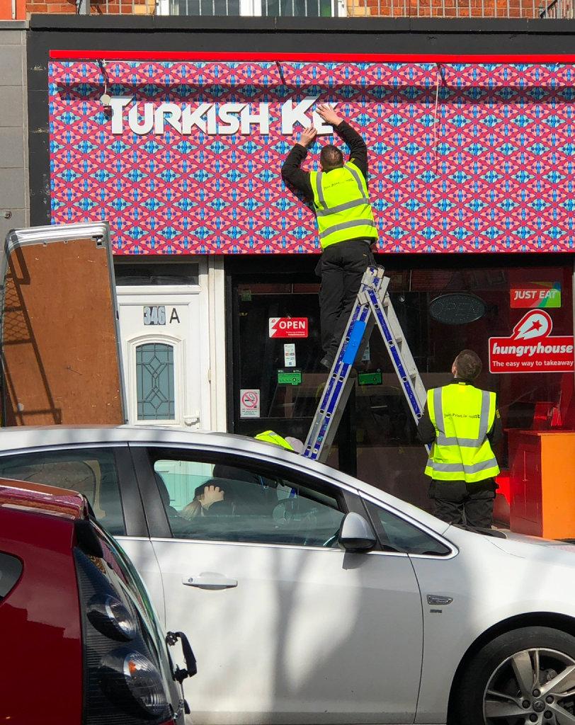 Wide shot of two workers in high-visibility vests on a ladder applying a large decorative wrap to the exterior sign of a shop.