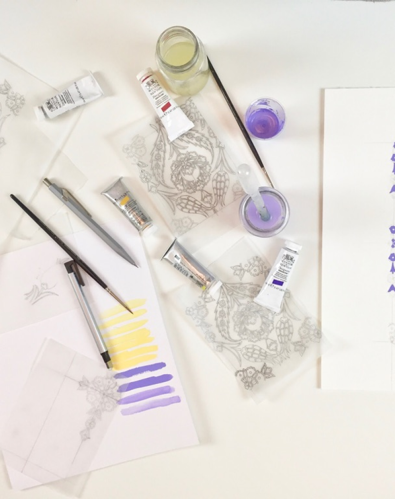 An overhead shot of an artist's desk with a compass, pencils, paint tubes, and a finished purple pattern on white paper.