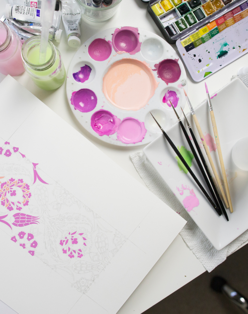 Top-down view of an art workspace featuring a round palette with various shades of pink and purple paint, brushes, and a floral sketch.