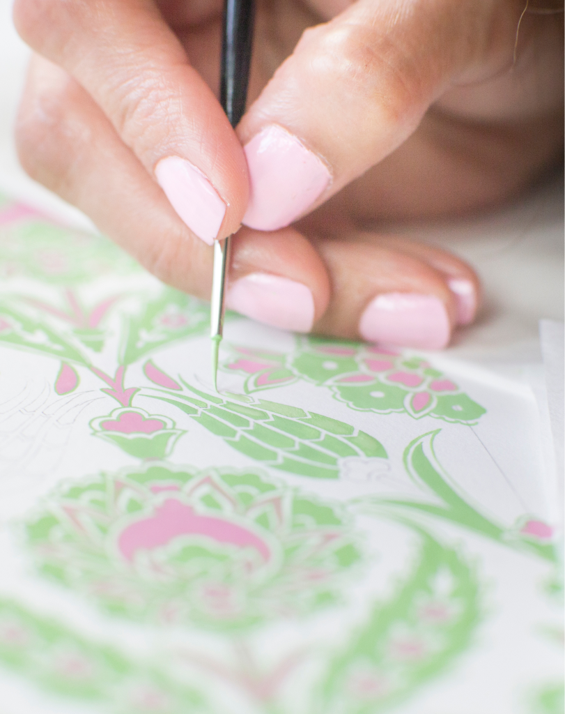 Close-up of a hand with pink nails using a fine-tipped brush to apply green paint to an intricate, traditional floral pattern.