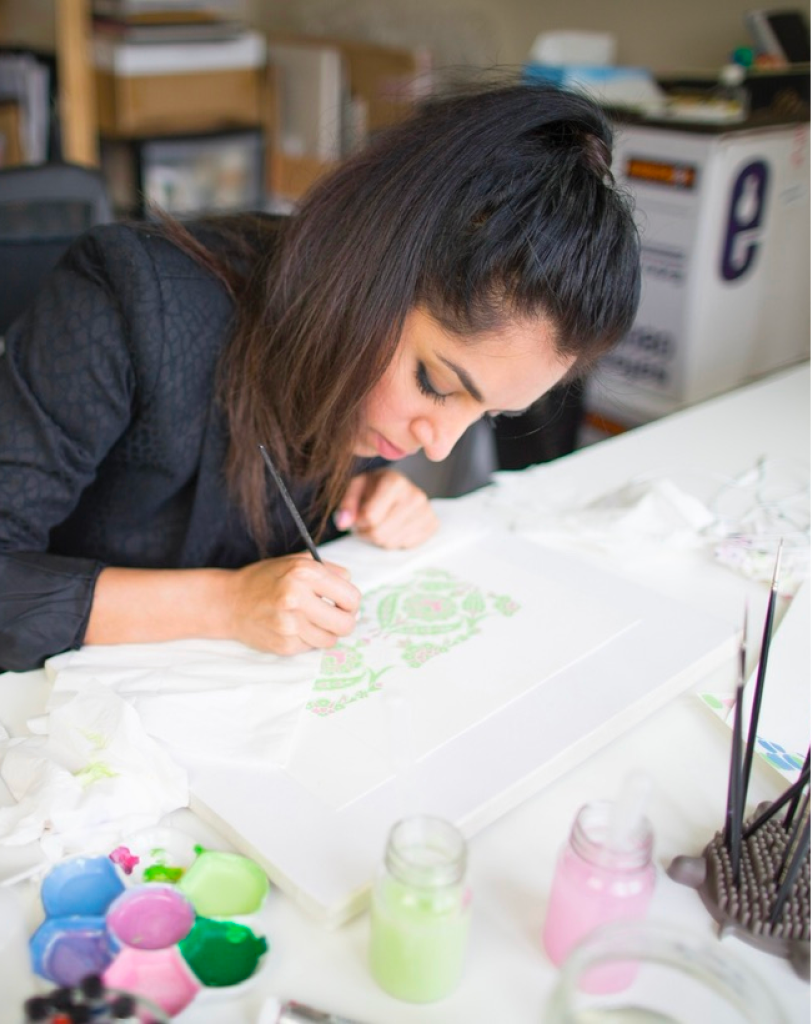 A woman with dark hair leaning over a desk, meticulously painting a large, detailed design with a fine brush in a bright studio.