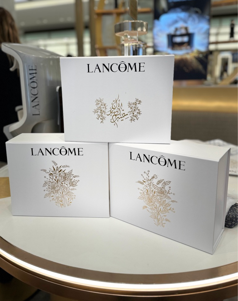 Three large white Lancôme gift boxes stacked in a pyramid shape on a table, each decorated with elegant gold floral motifs and one featuring gold Arabic calligraphy.