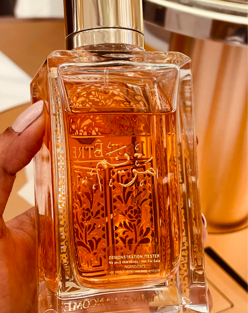 A close-up of a Maison Lancôme 'Peut-Être' perfume bottle held in a hand, showcasing the intricate gold lattice design and engraved Arabic script on the front of the bottle.