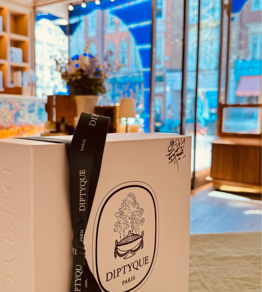 A vertical shot of a white Diptyque gift box standing upright, tied with a black ribbon. Small Arabic calligraphy is visible in the top right corner of the box, with a blurred view of a London street through the boutique window in the background.