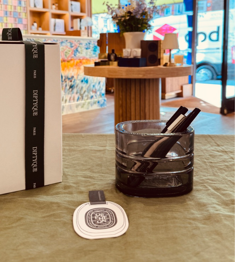A close-up of a green fabric tabletop featuring a Diptyque Orphéon fragrance tag and a smoky grey glass jar holding black pens. A white gift box with a black "Diptyque Paris" ribbon is partially visible on the left, set against a blurred boutique interior.