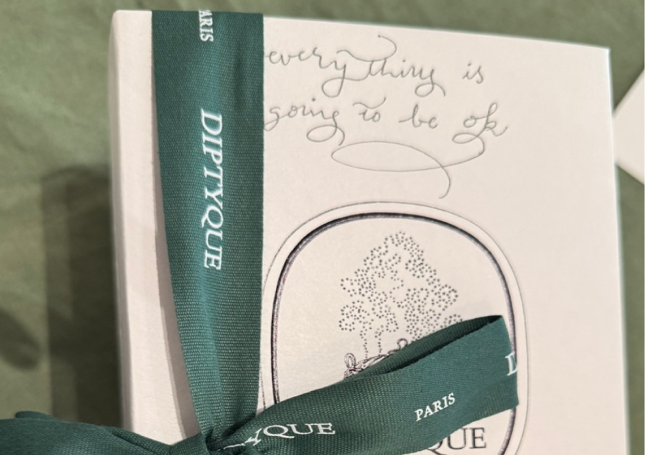 A close-up of a white gift box corner tied with a forest green Diptyque ribbon. Above the central logo, the phrase "everything is going to be ok" is delicately hand-written in light grey cursive script.