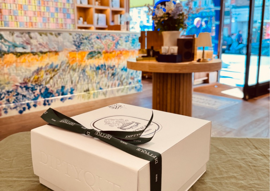 A white Diptyque gift box with an embossed logo and a black branded ribbon sits on a green fabric surface. The background features a brightly lit luxury boutique with a colorful impressionist-style tiled counter and wooden displays.