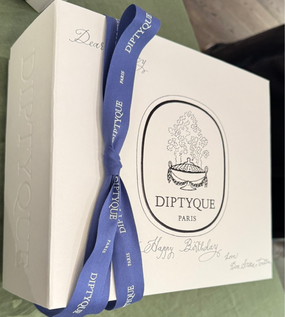 A top-down view of a white Diptyque gift box tied with a vibrant lavender-blue ribbon. Hand-lettered calligraphy on the box reads "Dear" at the top and "Happy Birthday Love Don, Stella & Caitlin" at the bottom.