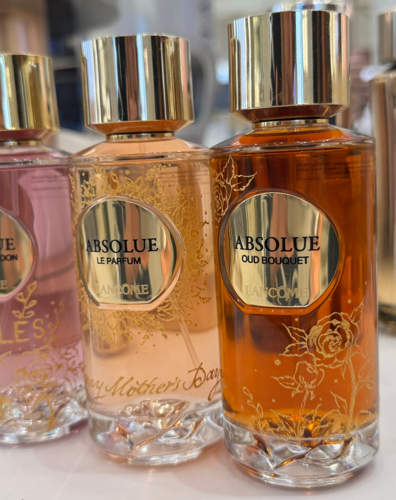 A row of four Lancôme Absolue perfume bottles—Le Parfum, Rose on the Moon, 1001 Roses, and Oud Bouquet—featuring gold caps and delicate floral gold leaf illustrations on the glass.