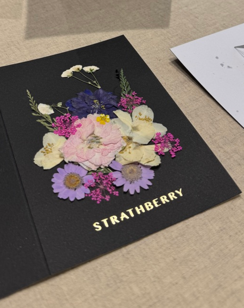 A flat-lay view of a black card being decorated with pressed purple, pink, and white flowers. Tweezers and floral scraps sit on the side.