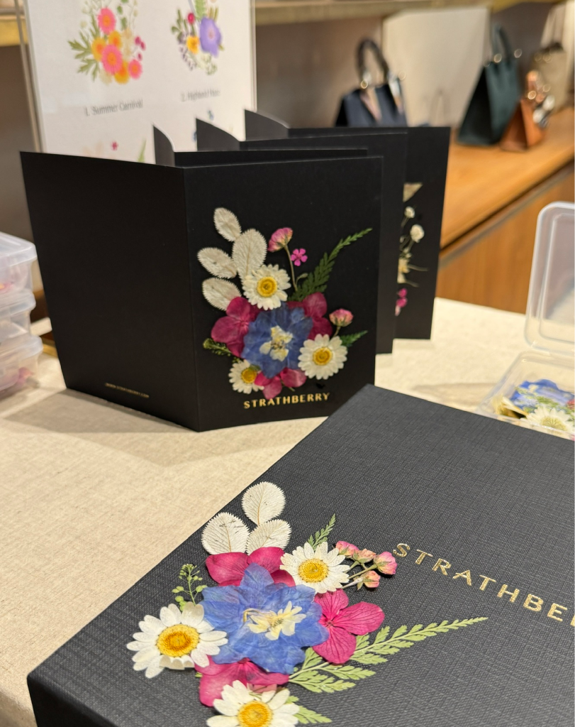 A close-up of a textured black card with a pressed flower arrangement of blue, pink, and white petals next to several finished cards.