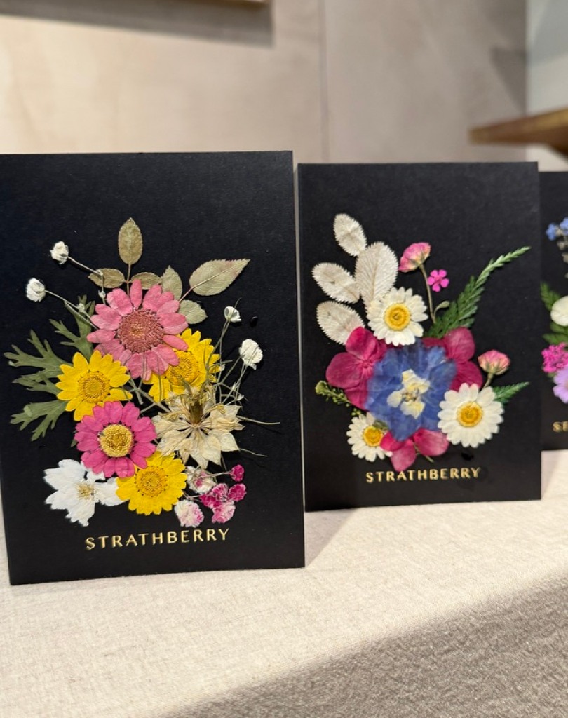 Two black Strathberry cards featuring intricate handmade bouquets of pressed pink, yellow, and white daisies with green foliage.