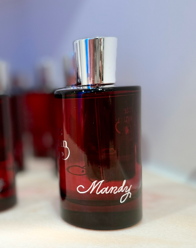 A vertical close-up of a personalized 'Juliette Has a Gun' fragrance bottle in deep red glass, with the name 'Mandy' engraved in white cursive at the bottom.