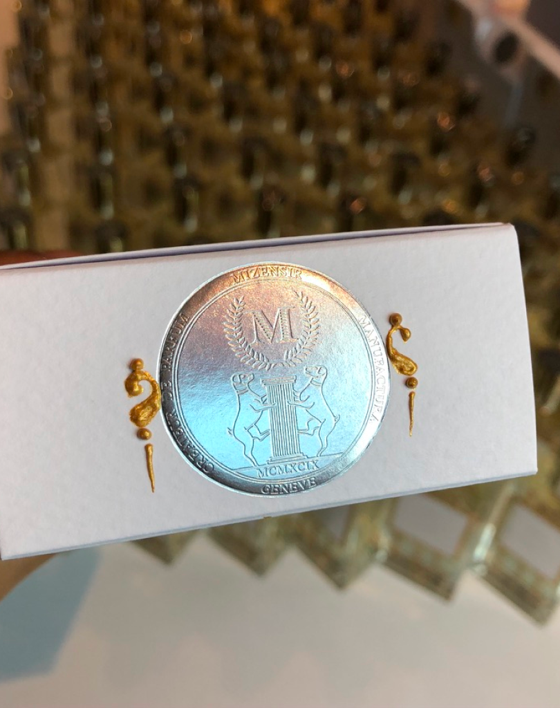 Close-up of a white textured Mizensir perfume box featuring a silver embossed circular seal with a laurel-wreathed "M," two dogs flanking a column, and gold wax-style accents on the sides.