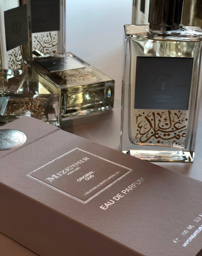 A lifestyle shot featuring a "Original Oud" perfume bottle next to its taupe-colored box. The bottle showcases intricate gold and silver hand-painted calligraphy details.