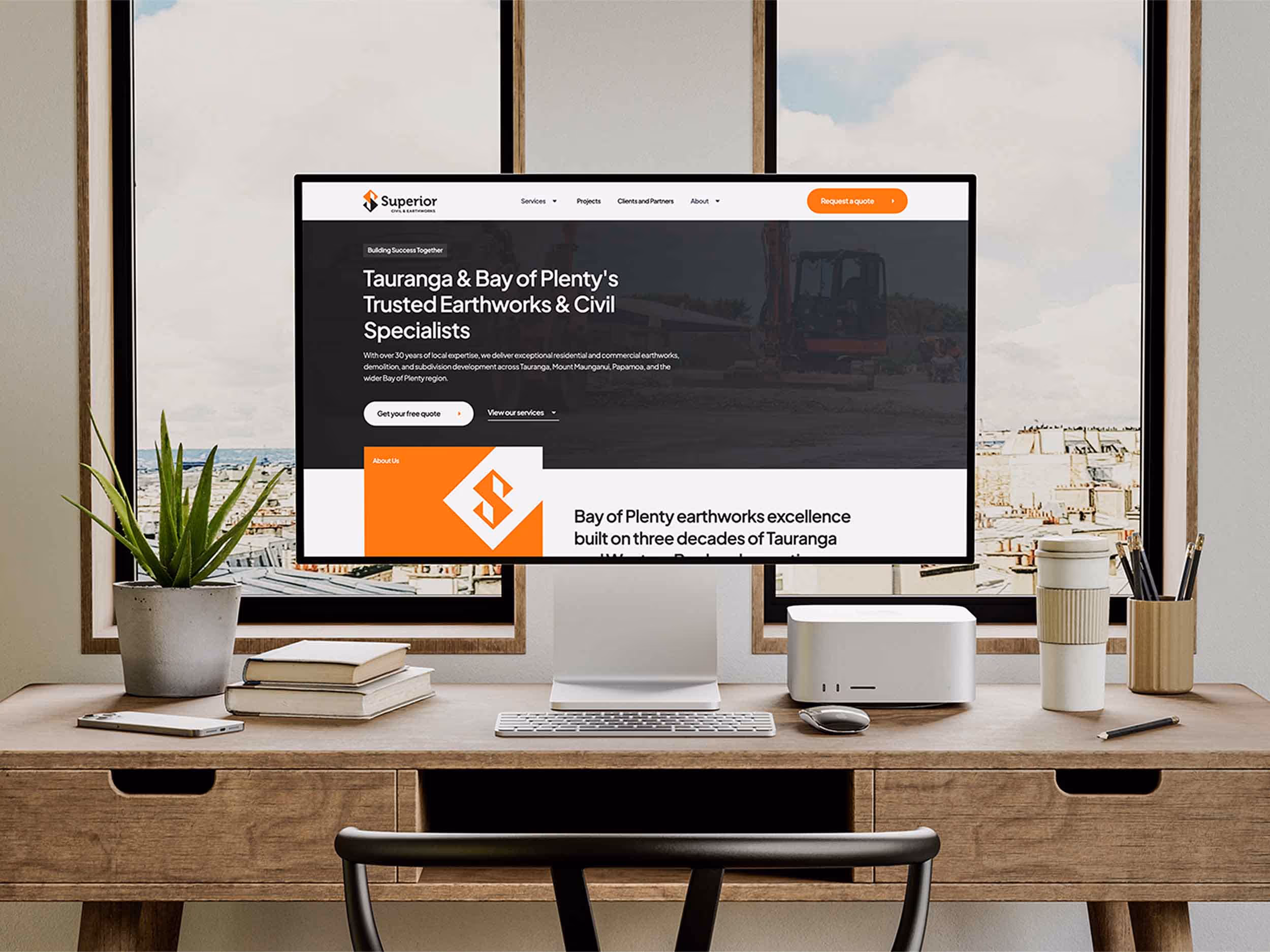 Modern home office with wooden desk holding a computer displaying a civil and earthworks company website, books, plant, coffee cup, and stationery, with large windows in the background.