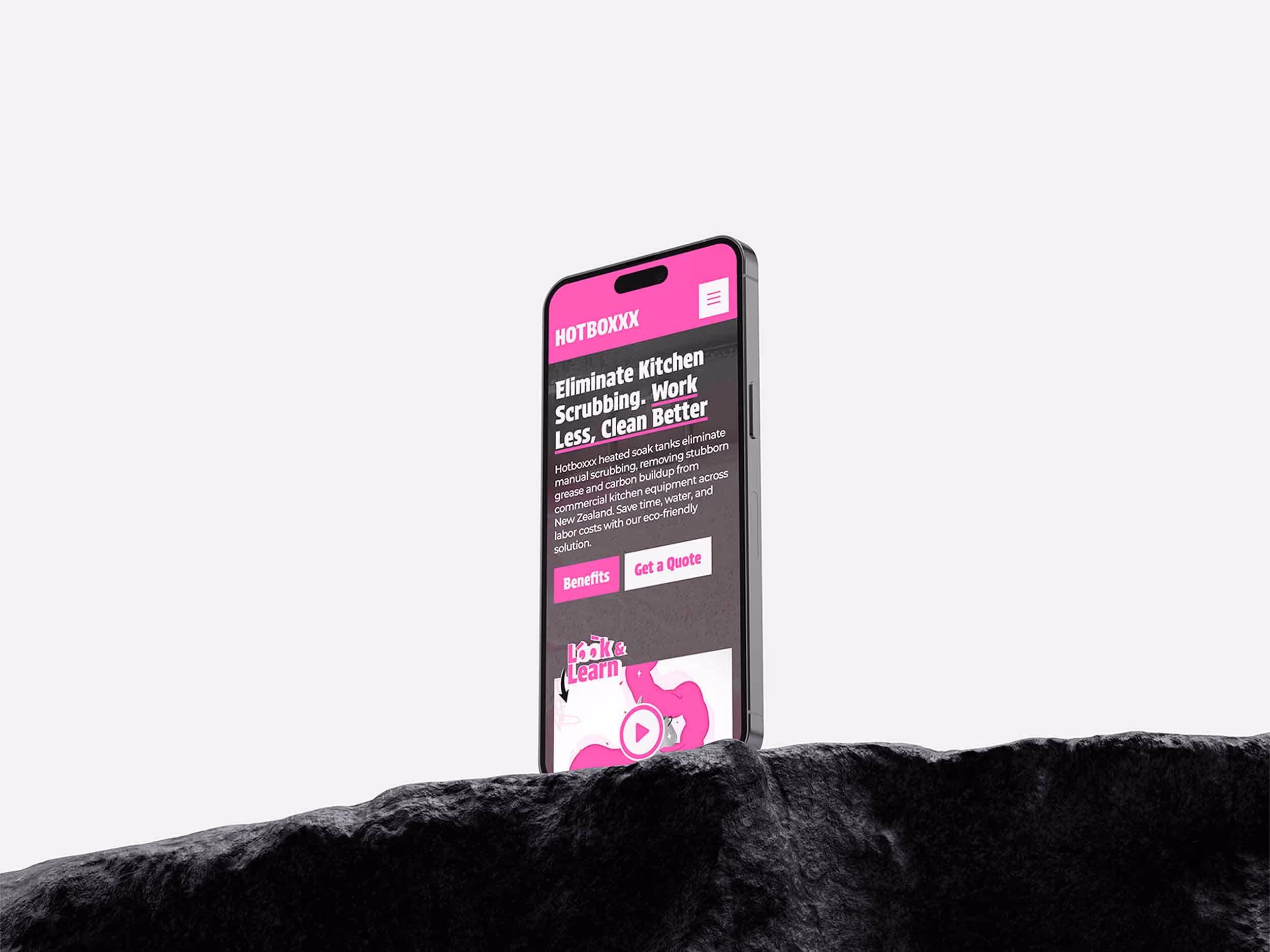 Smartphone displaying HOTBOXXX website about eliminating kitchen scrubbing with heated soak tanks, positioned behind a black rocky surface.