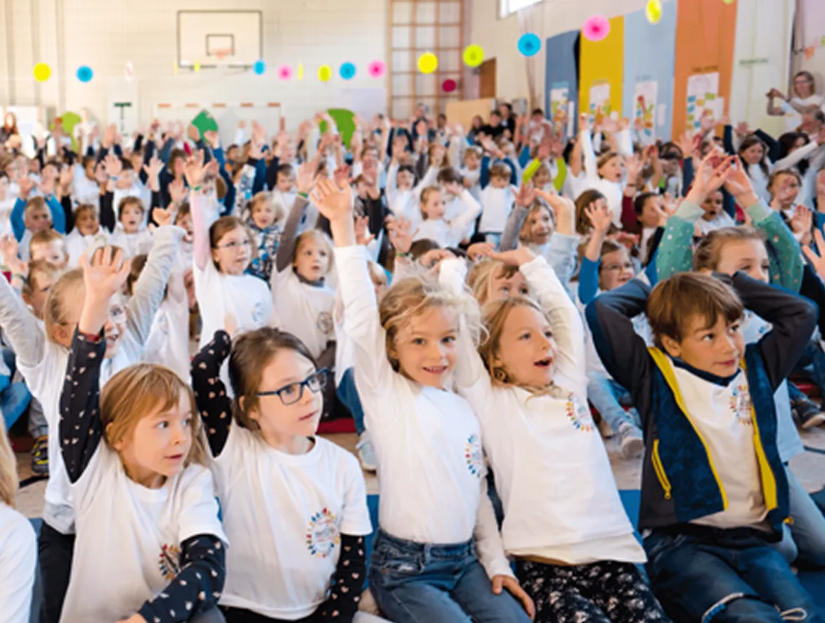 Viele Kinder in weißen T-Shirts sitzen in einer Turnhalle und heben ihre Hände begeistert.