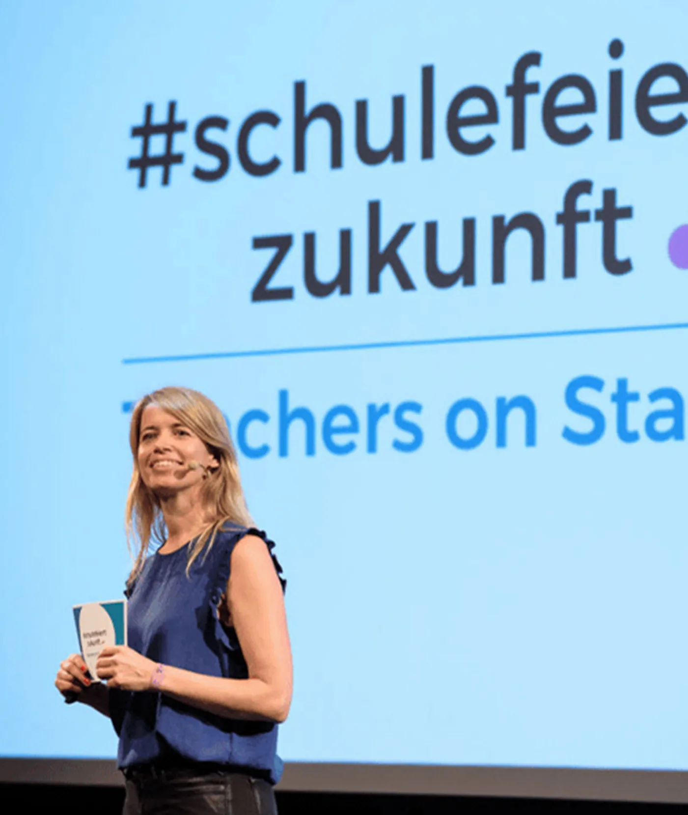 Eine Frau mit blauer Bluse hält Karten und spricht vor einer großen blauen Leinwand mit der Aufschrift „#schulefeier zukunft“.