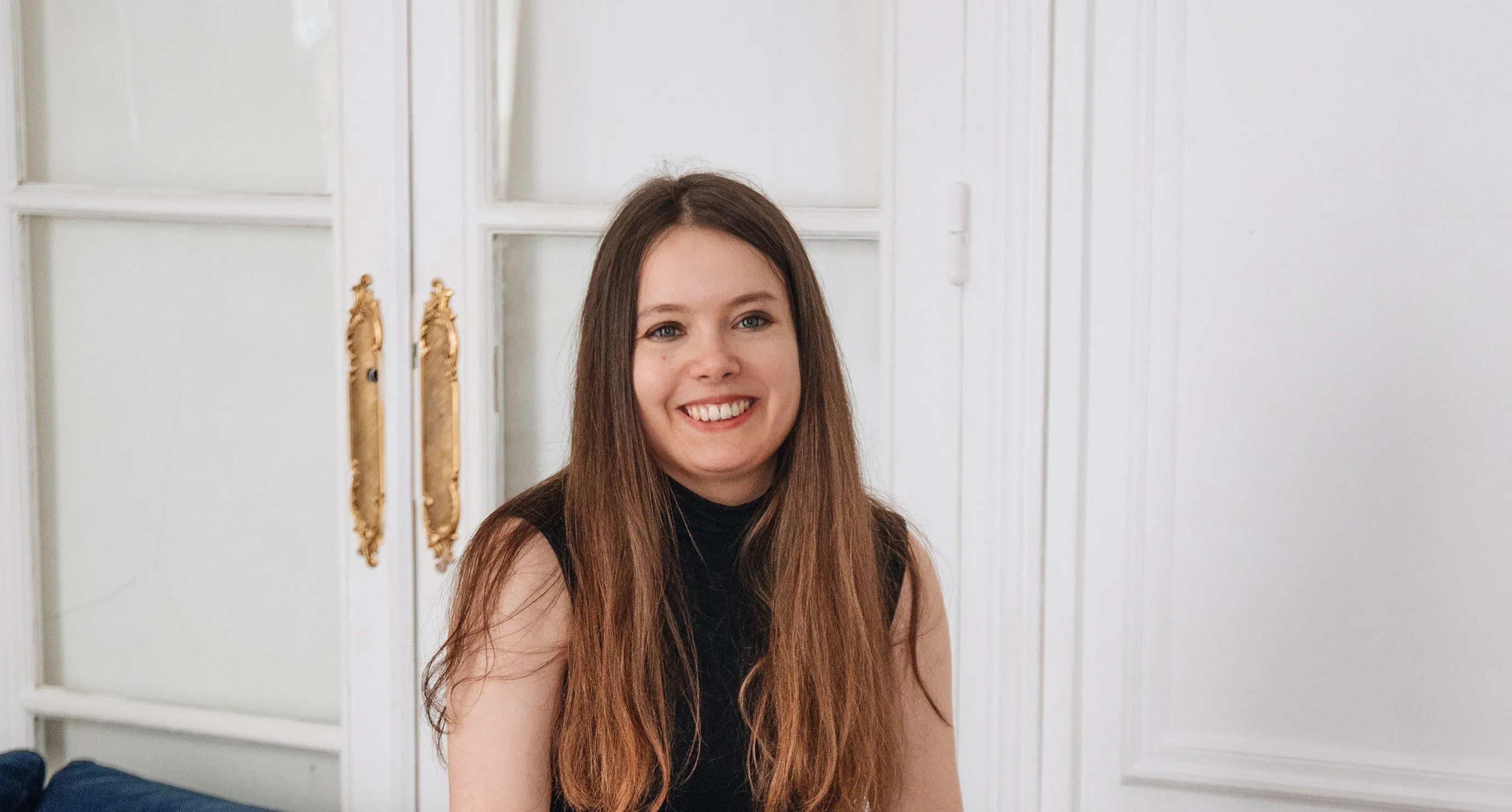 Smiling young woman with long brown hair wearing a black sleeveless top, seated in front of white paneled doors with gold handles.