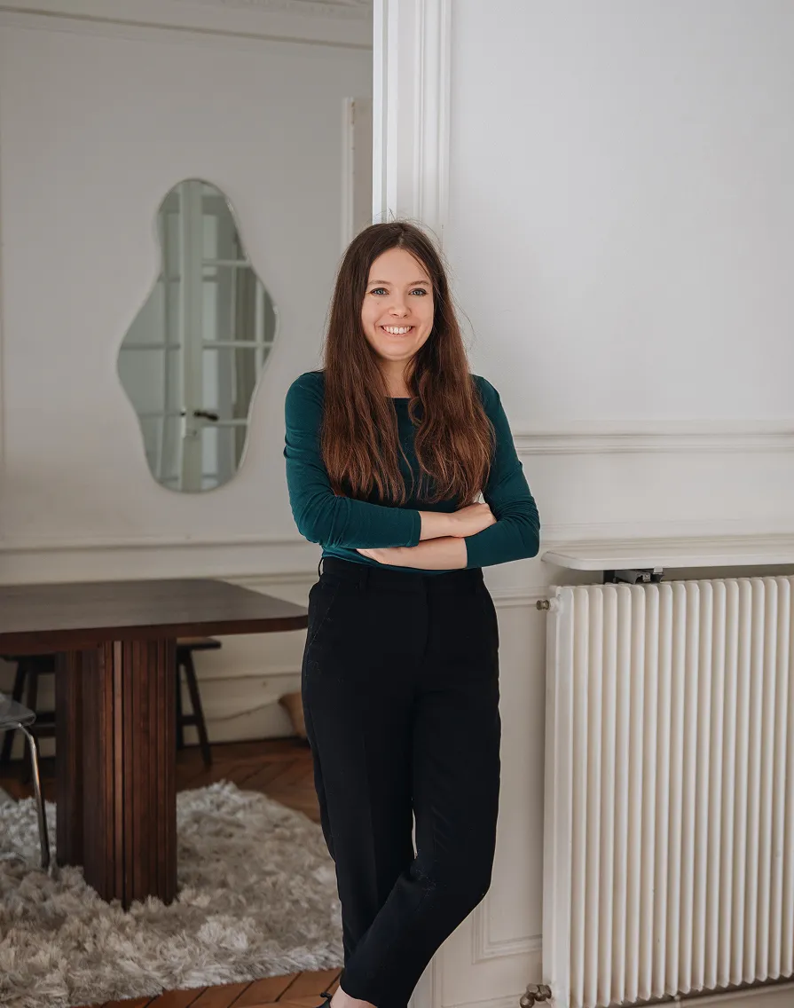Smiling woman with long brown hair wearing a dark green long-sleeve shirt and black pants, standing with arms crossed in a room with white walls and a radiator.