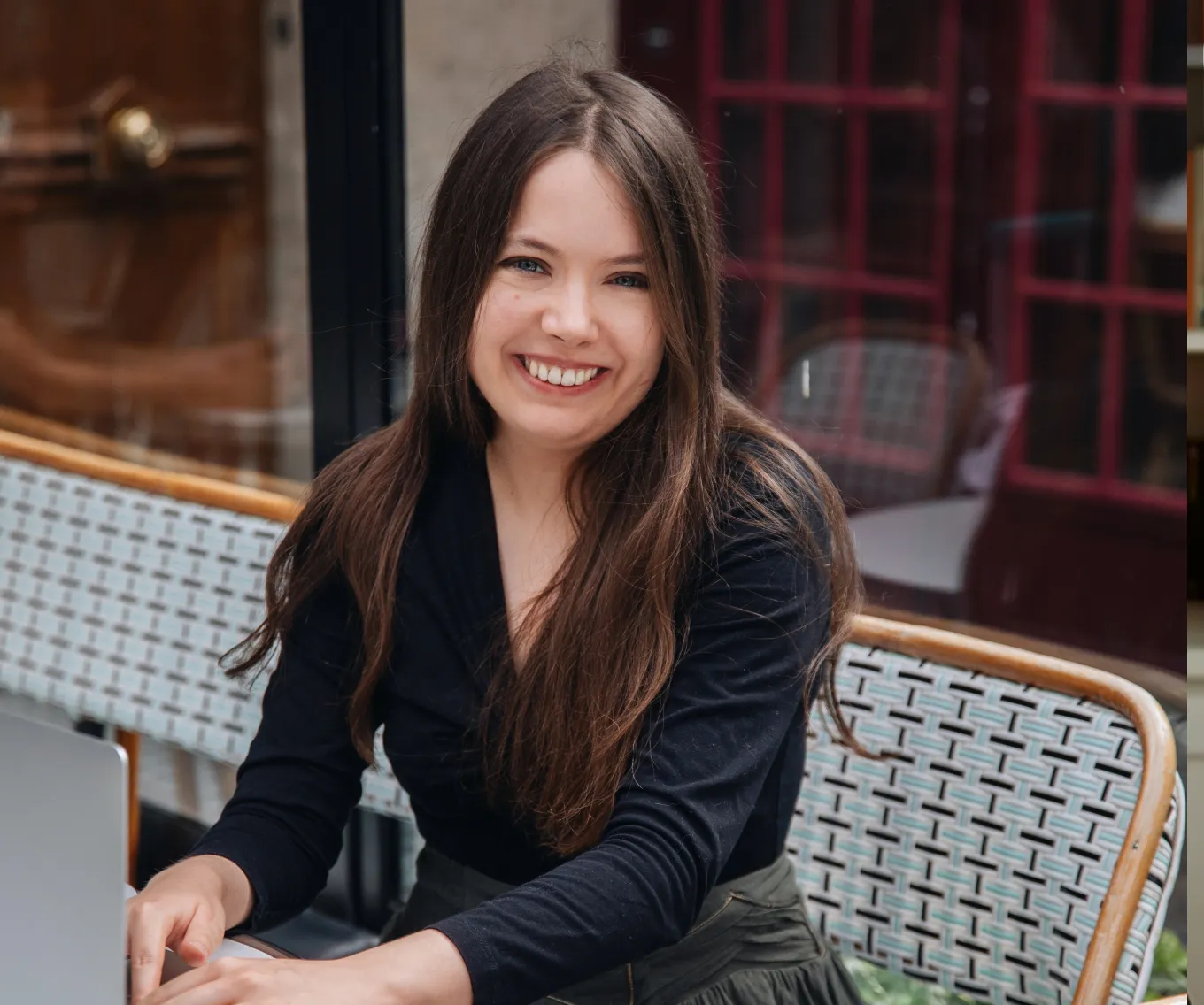 Smiling woman with long brown hair sitting at an outdoor table working on a laptop.