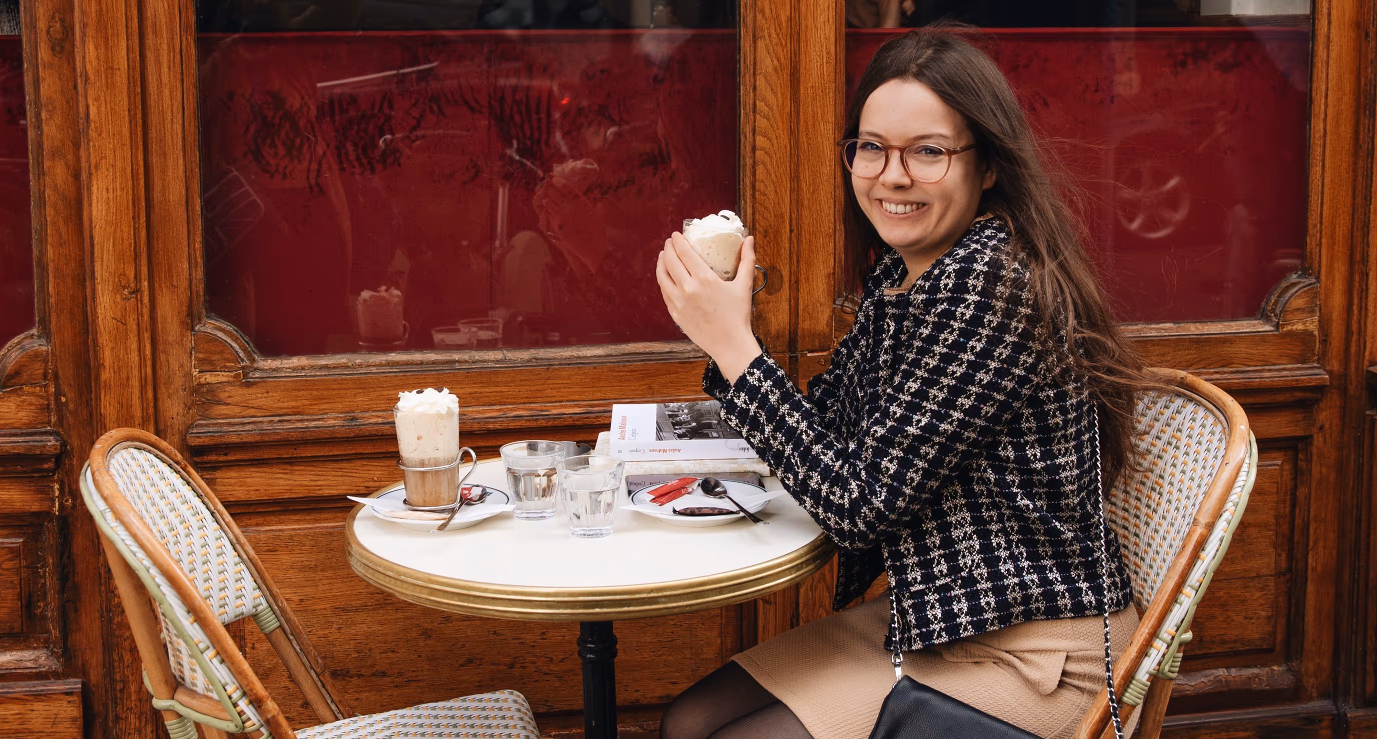 Smiling woman with glasses sitting at a small café table holding a cup of coffee topped with whipped cream.
