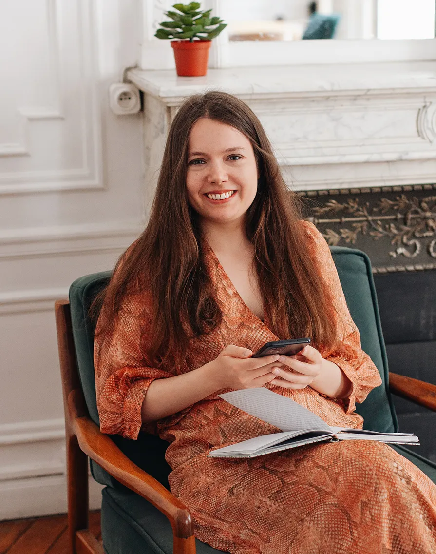 Smiling woman with long brown hair in a patterned orange dress sitting on a green chair holding a smartphone with an open notebook on her lap.