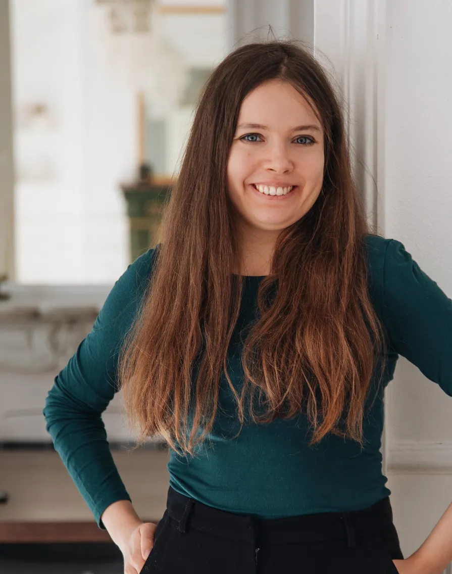 Smiling woman with long brown hair wearing a teal long-sleeve shirt and black pants leaning against a white wall.