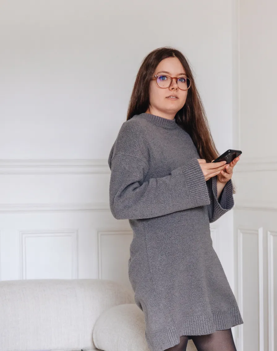 Woman with long brown hair and glasses wearing a gray sweater dress, holding a smartphone indoors.