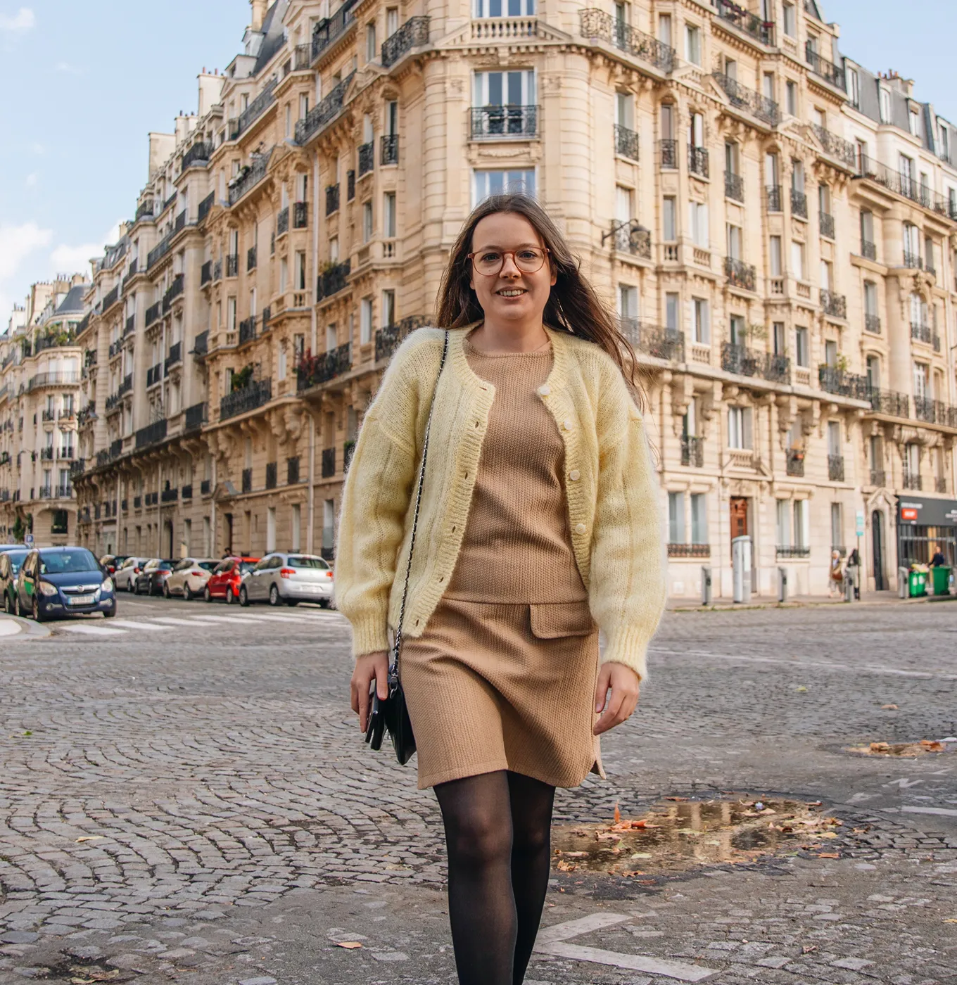 Young woman in a beige dress and yellow cardigan walking on a cobblestone street with historic buildings in the background.