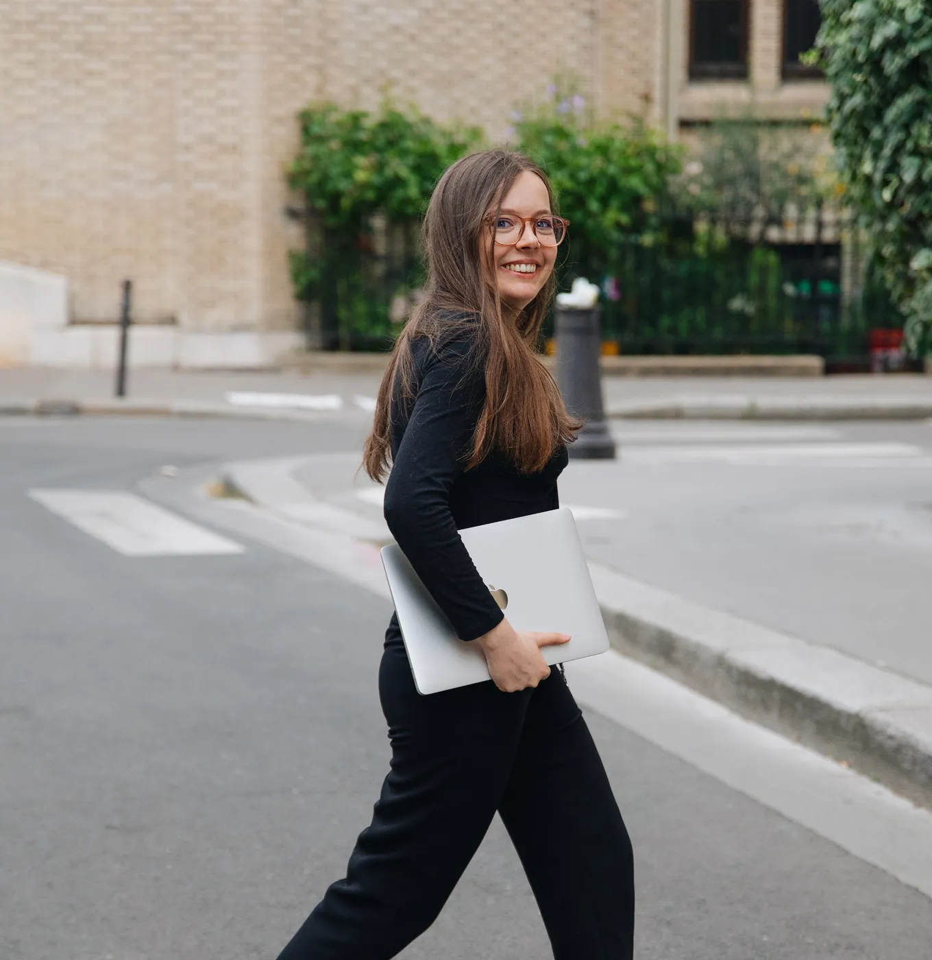 Smiling woman with glasses walking outdoors carrying a closed laptop under her arm.