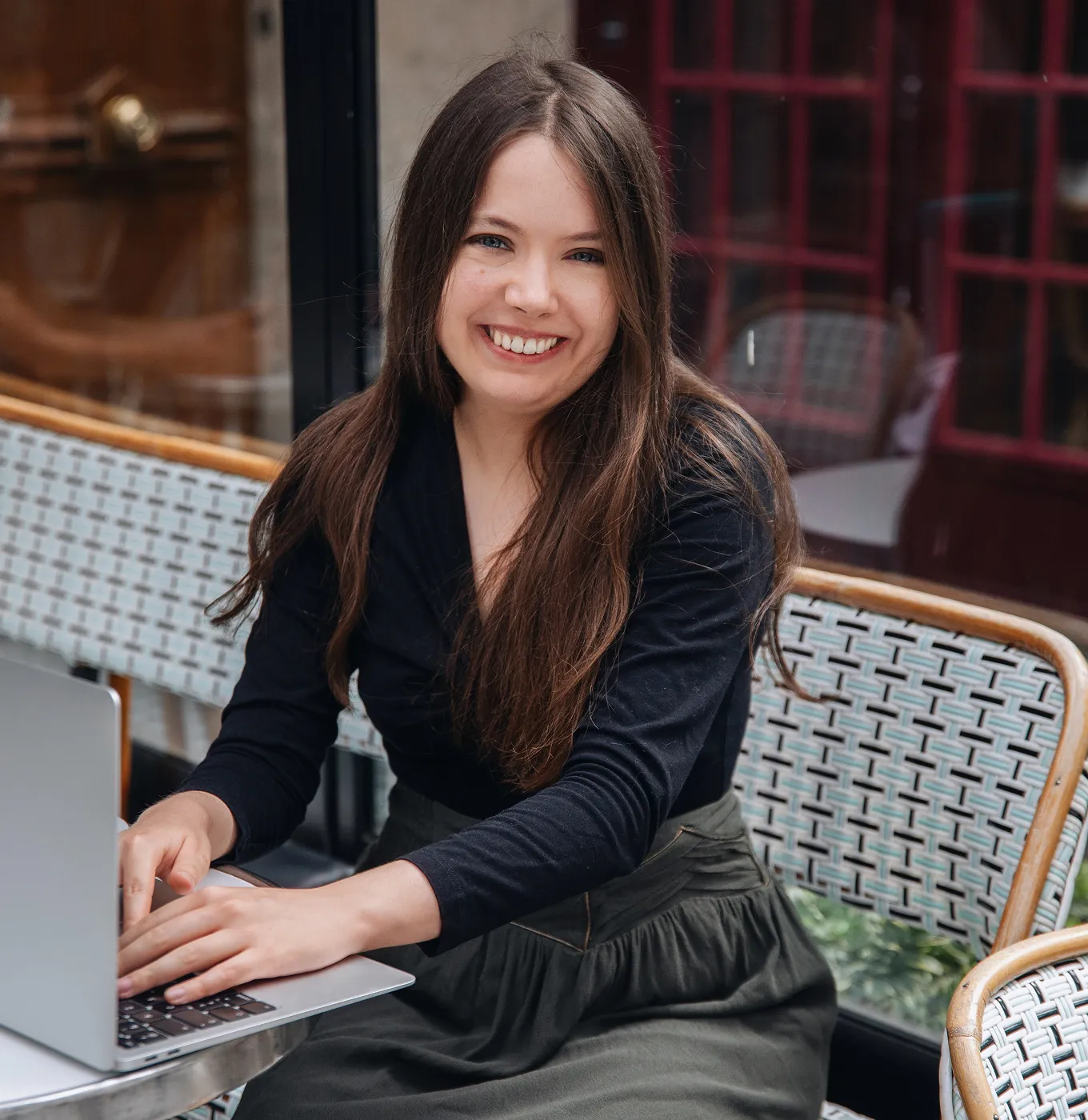 Smiling woman with long brown hair typing on a laptop at a cafe table.