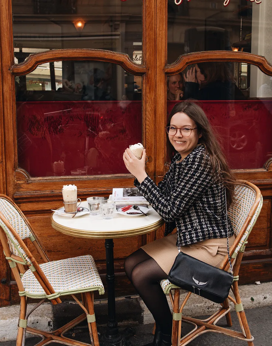 Smiling woman with glasses sitting at an outdoor café table holding a cup topped with whipped cream.