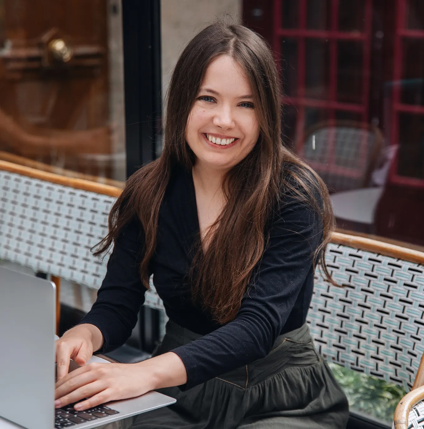 Smiling woman with long brown hair sitting outdoors using a laptop on a woven chair.