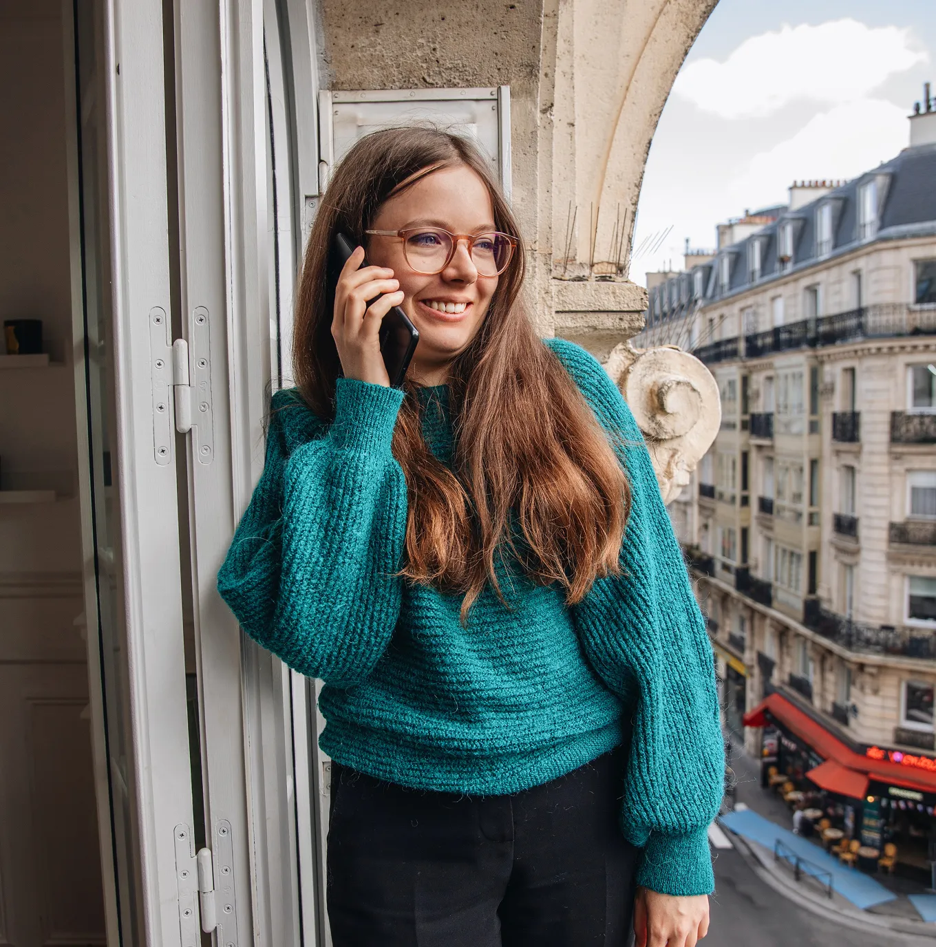 Smiling young woman with glasses and long brown hair in a teal sweater talking on a smartphone by an open window overlooking a city street with classic buildings.