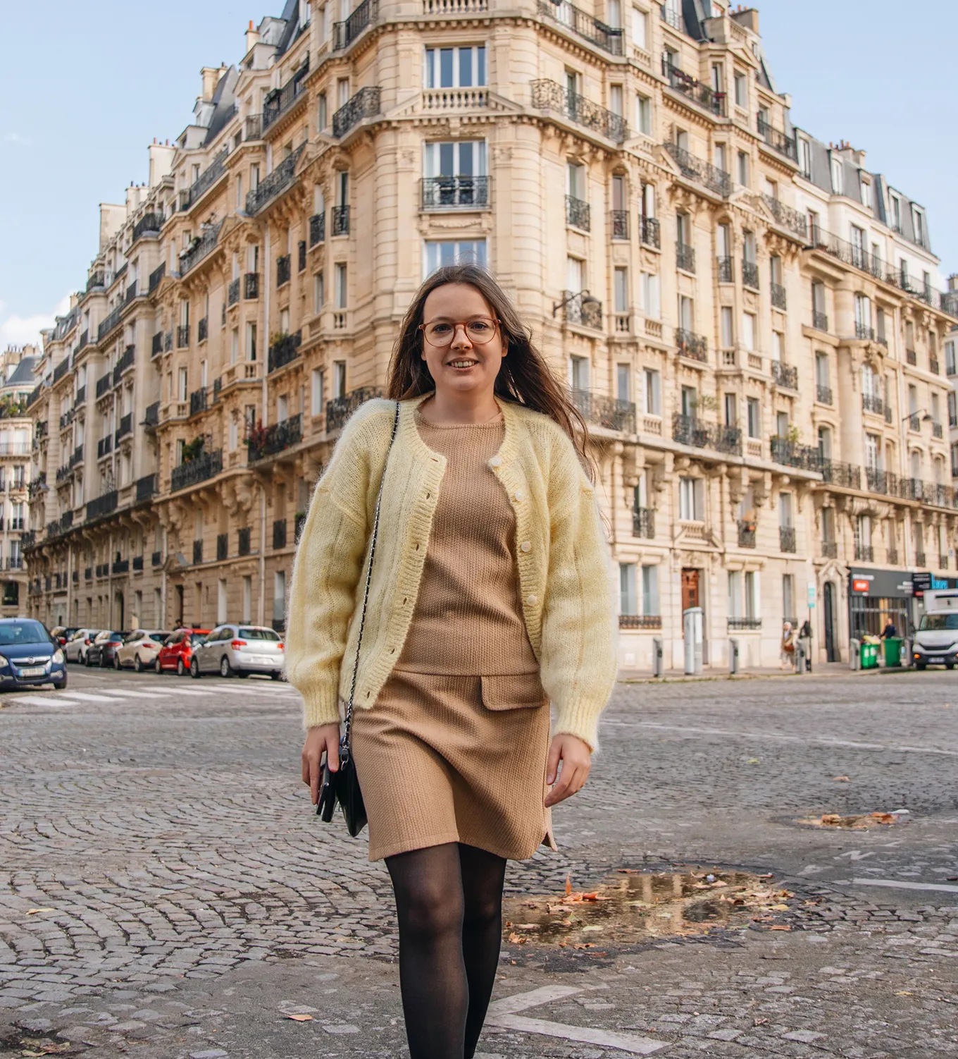 Young woman wearing glasses and beige outfit with yellow cardigan walking on a cobblestone street in front of classic Parisian buildings.