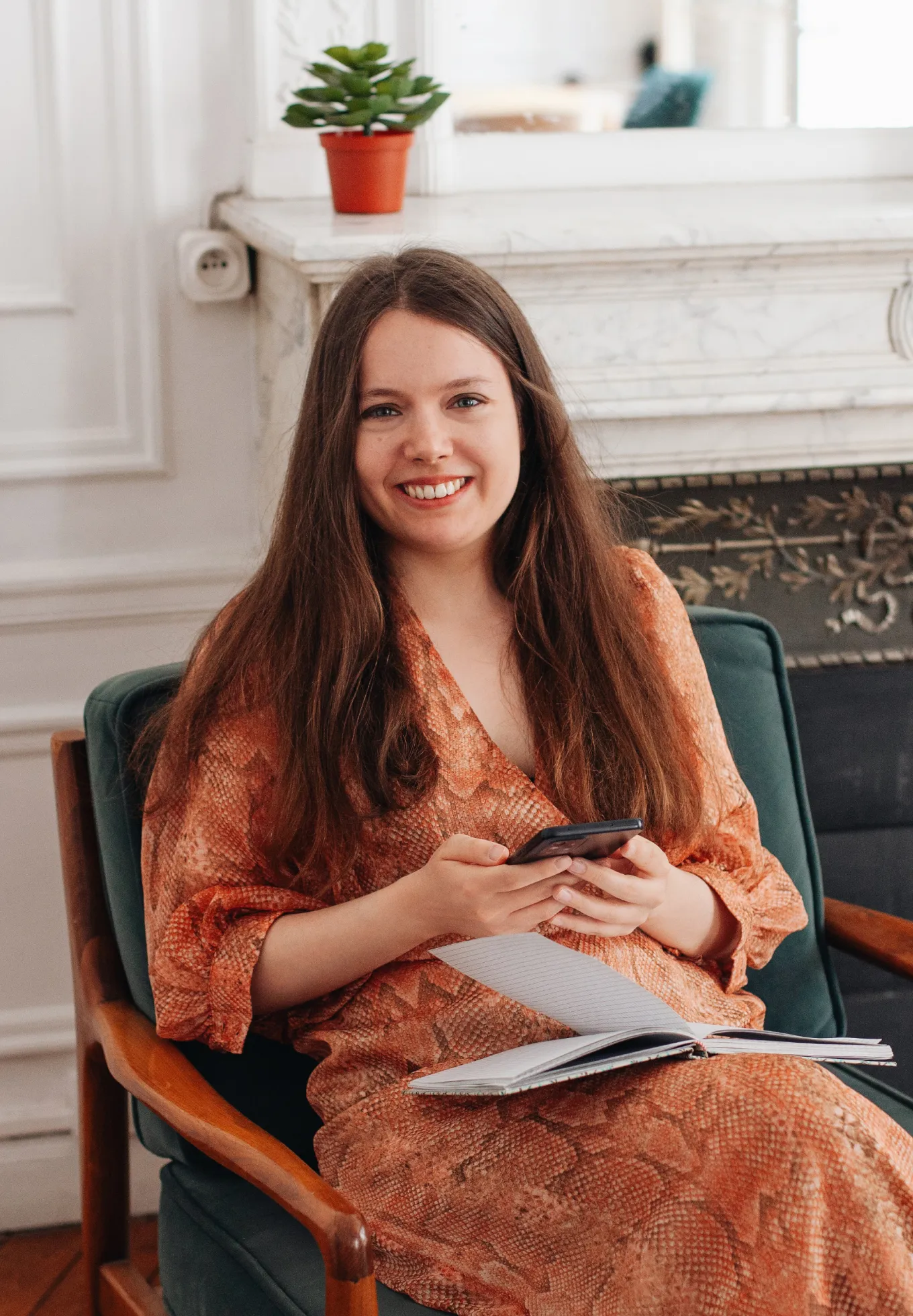 Jeune femme souriante assise sur un fauteuil vert, tenant un téléphone et un carnet ouvert.