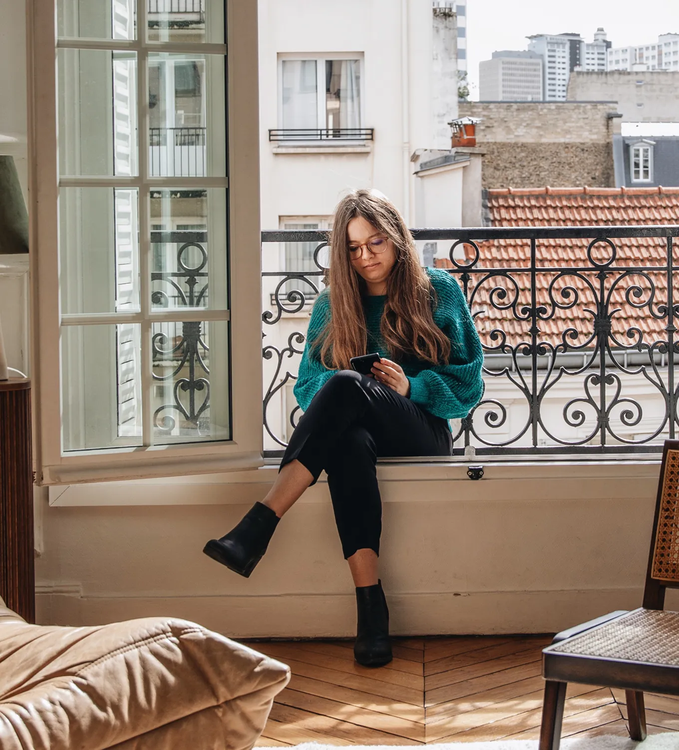 Jeune femme aux cheveux longs assise sur le rebord d'une fenêtre ouverte, regardant son téléphone, avec un pull turquoise et des bottines noires dans un appartement urbain.