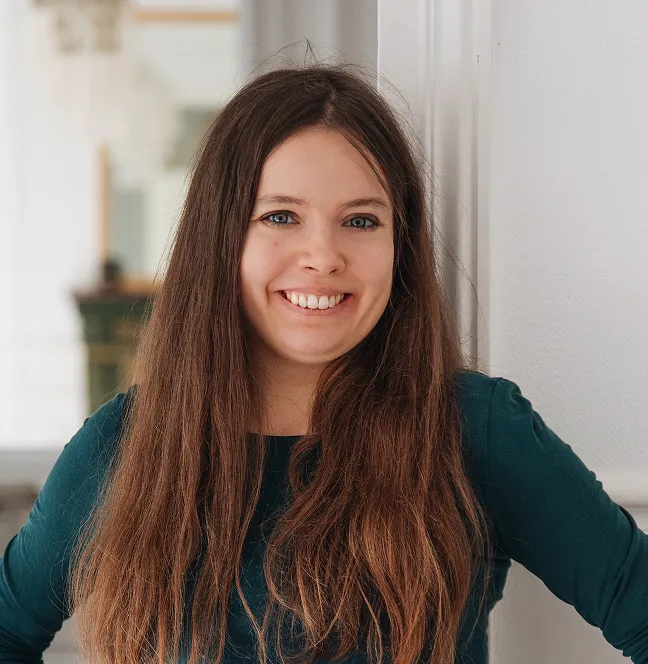 Jeune femme aux cheveux longs et bruns souriant, appuyée contre un mur blanc dans un intérieur lumineux.