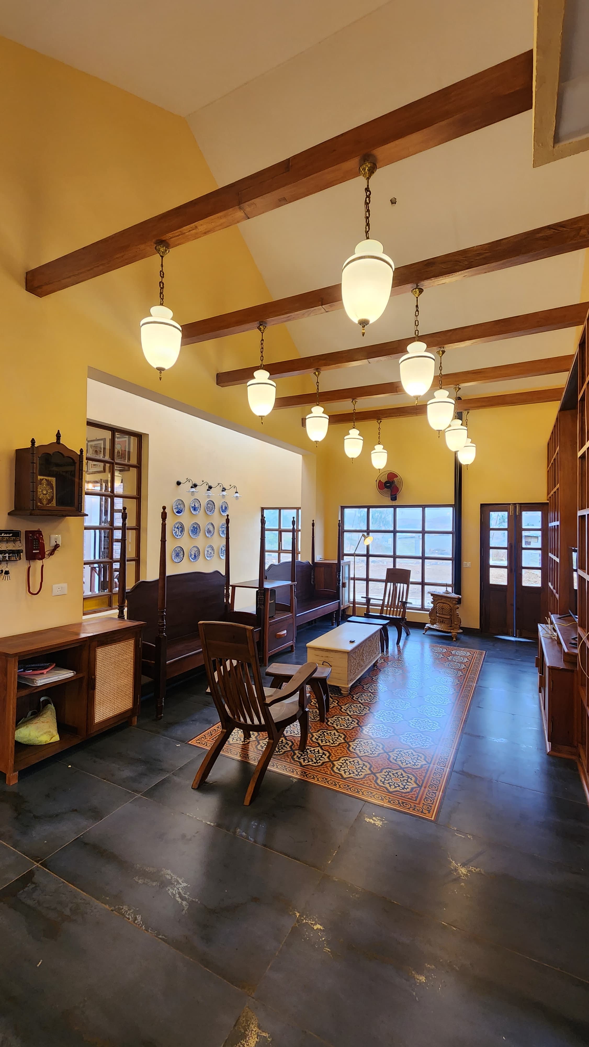Bright living room with wooden ceiling beams, hanging pendant lights, dark tile floor, and vintage wooden furniture around a decorative rug.