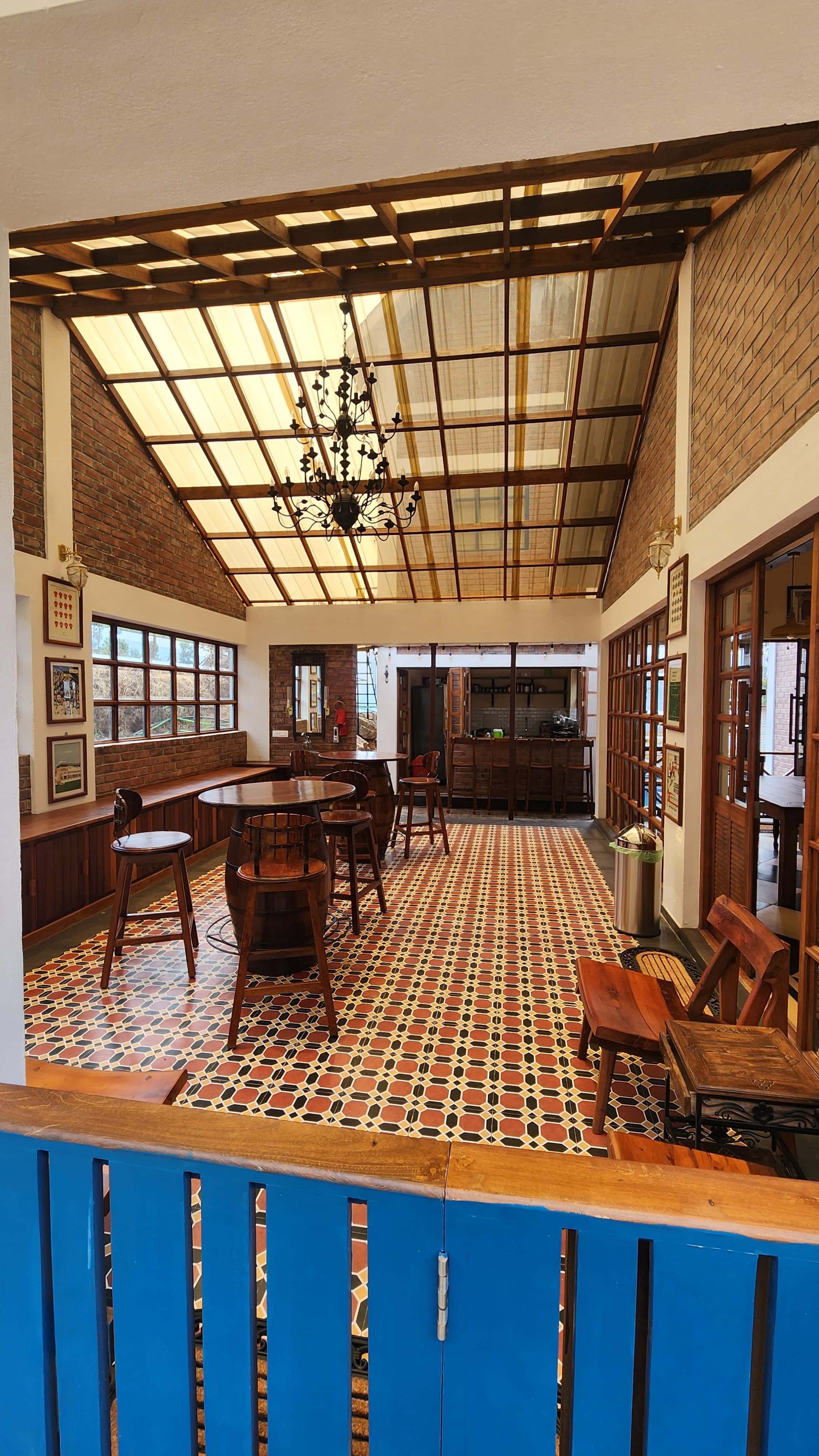 Indoor seating area with wooden tables and chairs, patterned tile floor, brick walls, and a transparent roof with a chandelier.