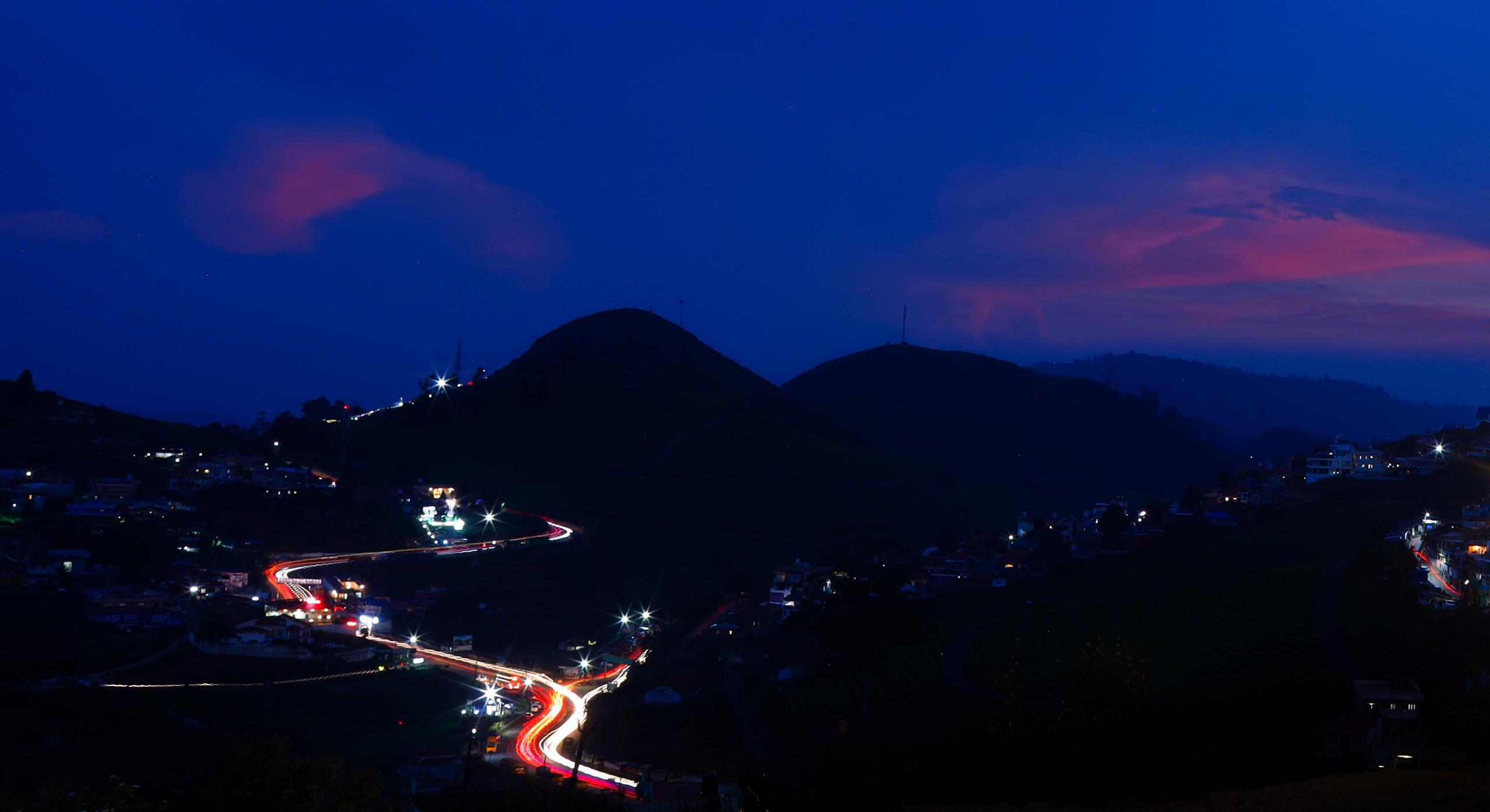 Winding road with car light trails through a mountainous village at dusk under a blue and purple sky.