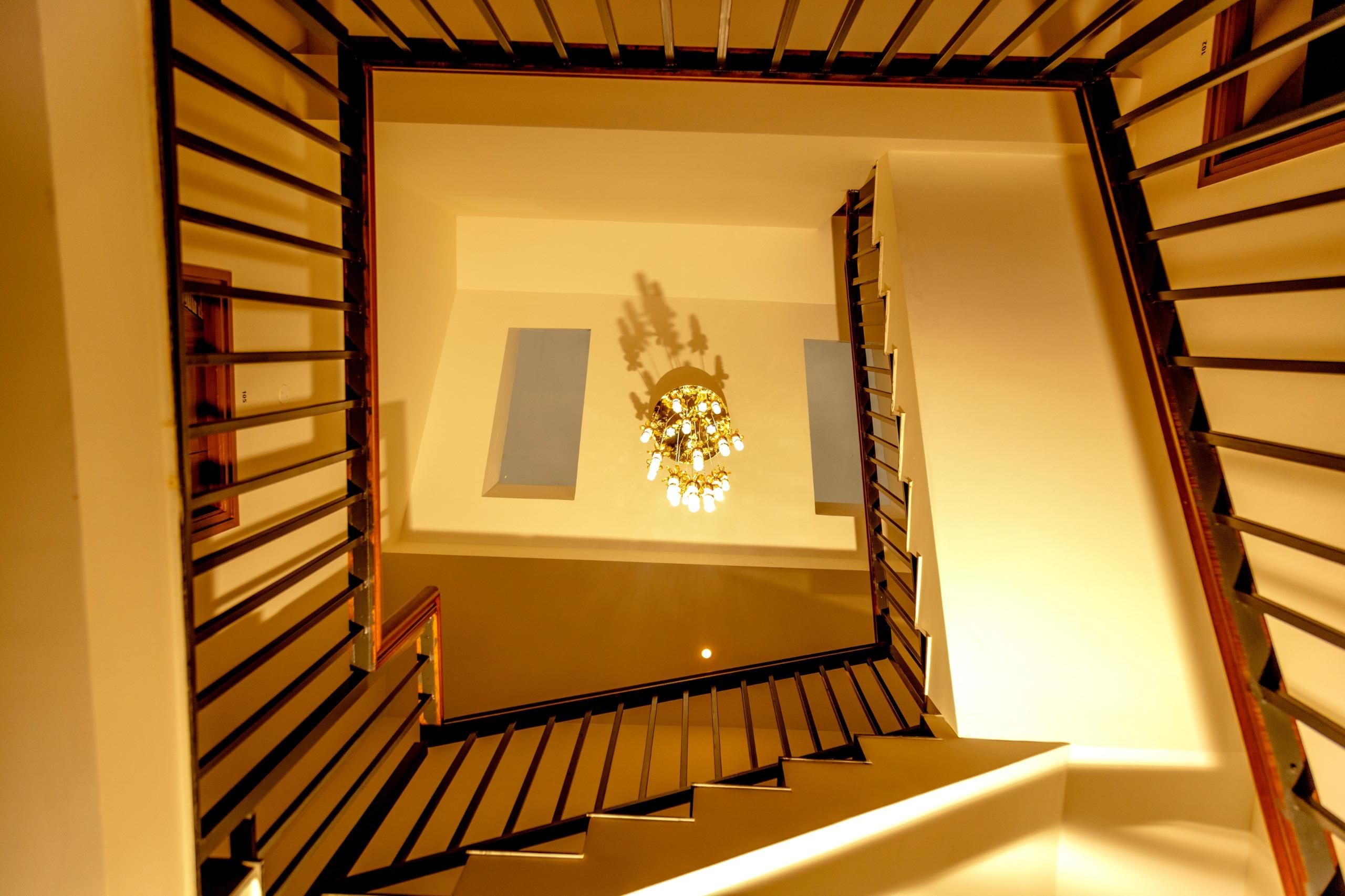 View looking up a square stairwell with black railings and a chandelier hanging from the ceiling.