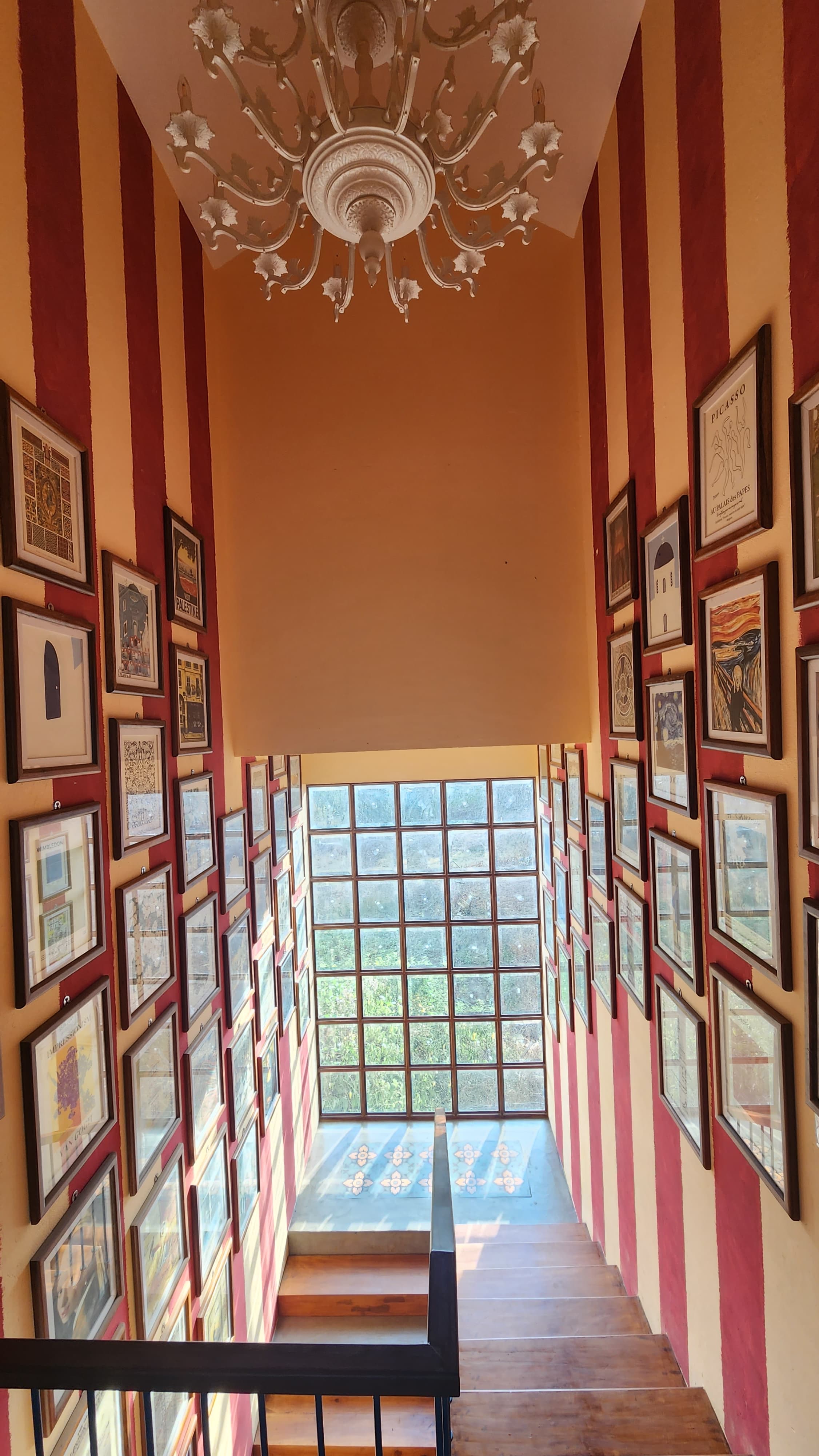 Staircase with wood steps and walls painted in vertical red and beige stripes, decorated with numerous framed artworks and a large window at the bottom.