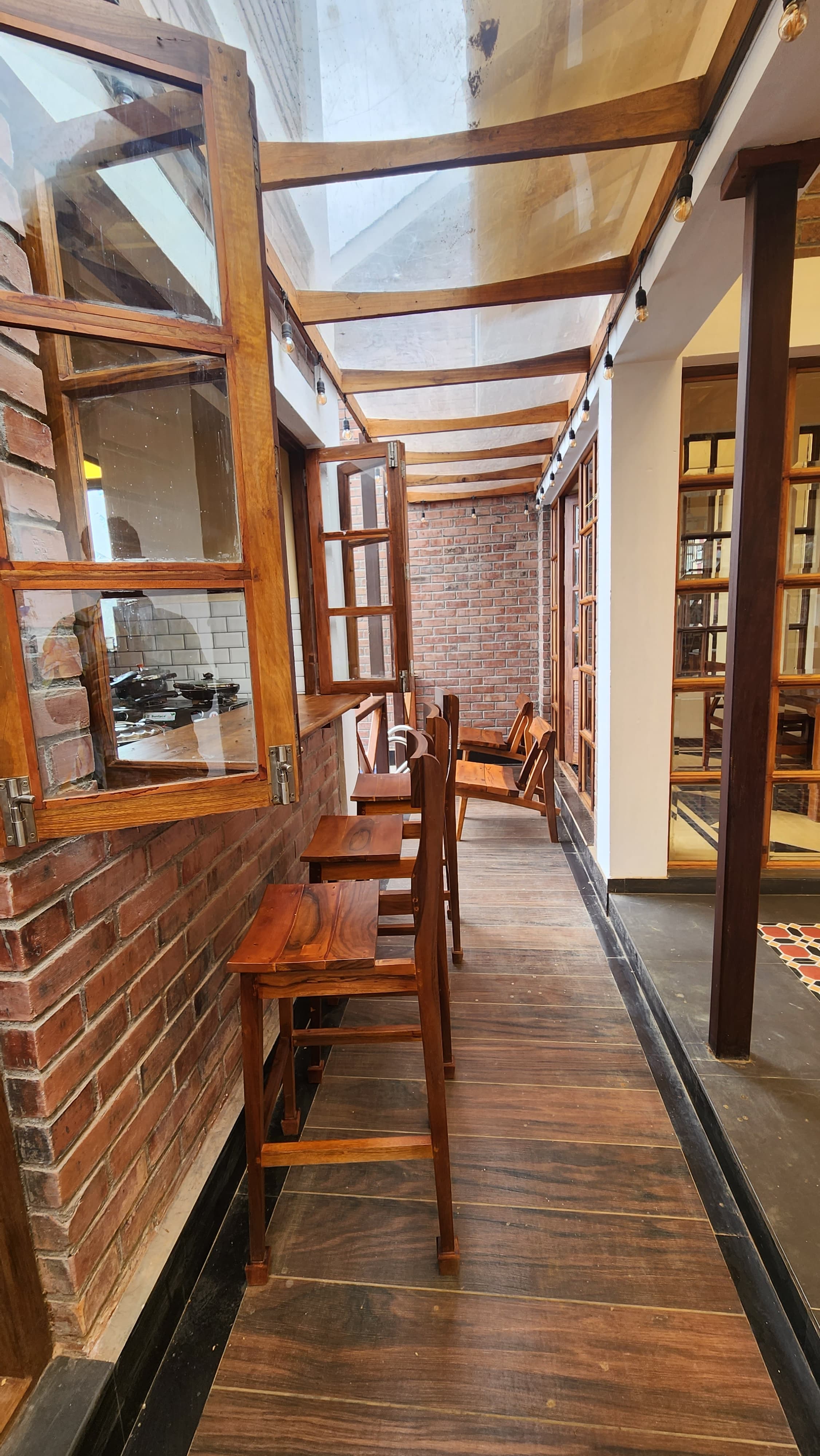 Narrow indoor seating area with wooden chairs and stools along brick walls and windows under a transparent roof with hanging lights.