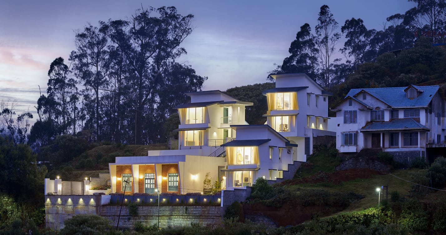 Modern hillside house with illuminated large windows surrounded by trees at dusk.